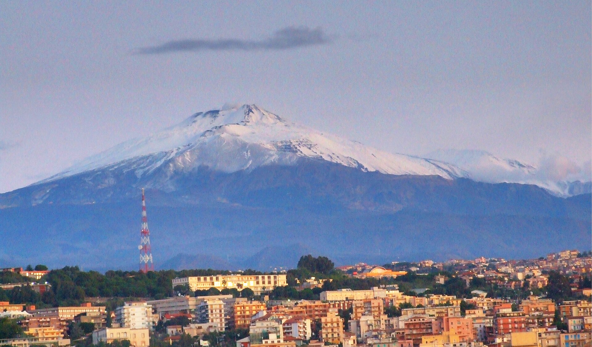 Cima innevata dell’Etna vista da una città in primo piano, con un’antenna rossa e bianca.