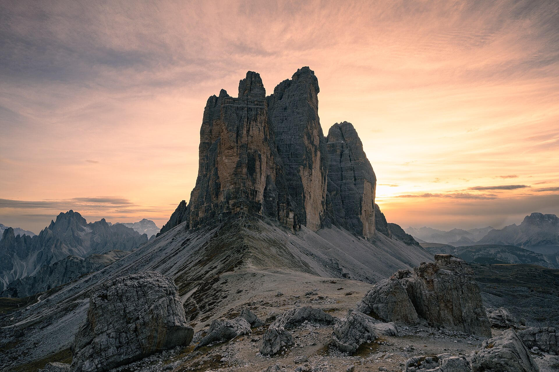 Tre Cime di Lavaredo al tramonto, tre guglie di dolomia su ghiaione, cielo arancio.